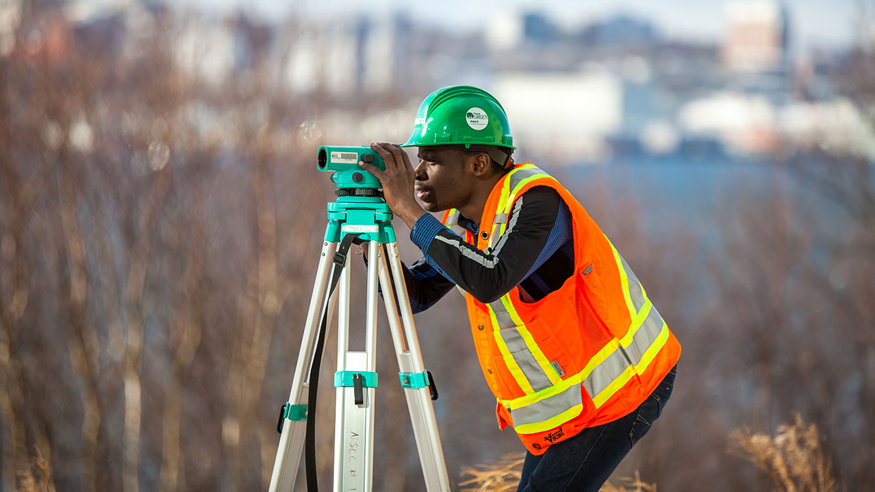 A student in a safety vest and hard hat bends and looks through a piece of surveying equipment.
