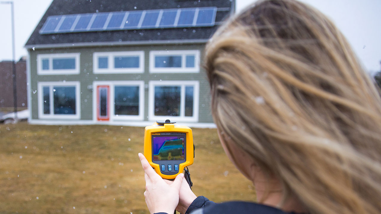 A person stands outdoors in front of a home and looks at the screen of a heat imaging tool.
