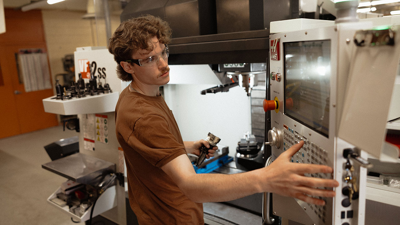 A student with safety glasses adjusts part of a machine in a workshop setting.