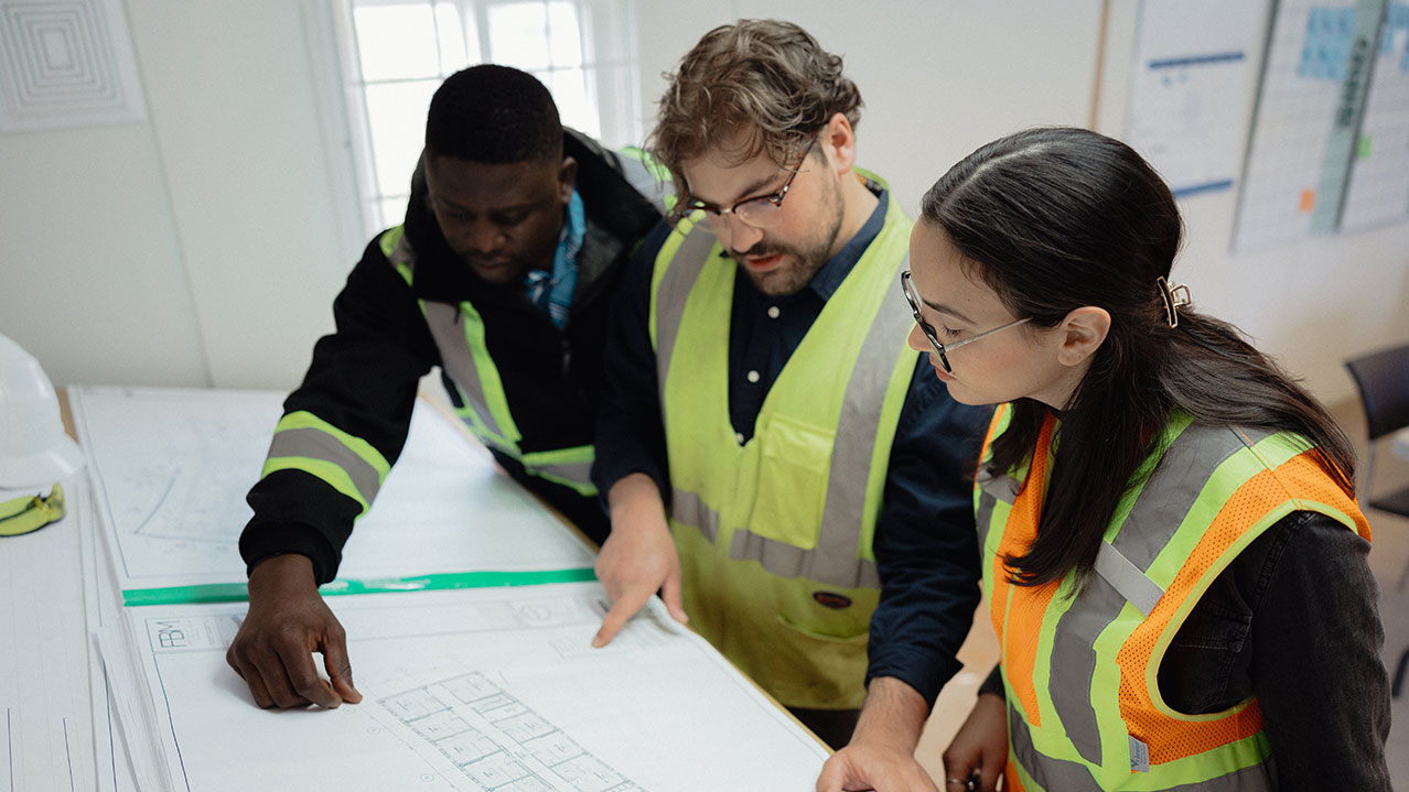 Three students in safety vests look at blue prints on a table.