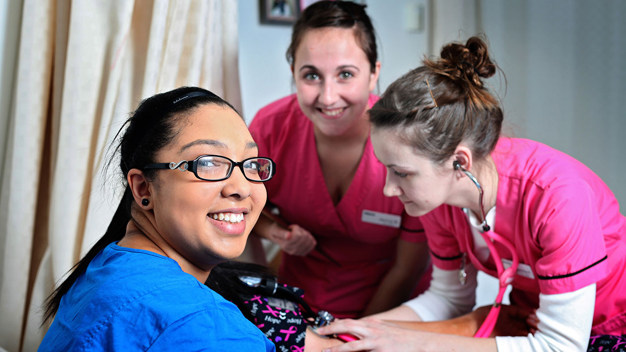 Three students in brightly coloured scrubs practise taking blood pressure readings.