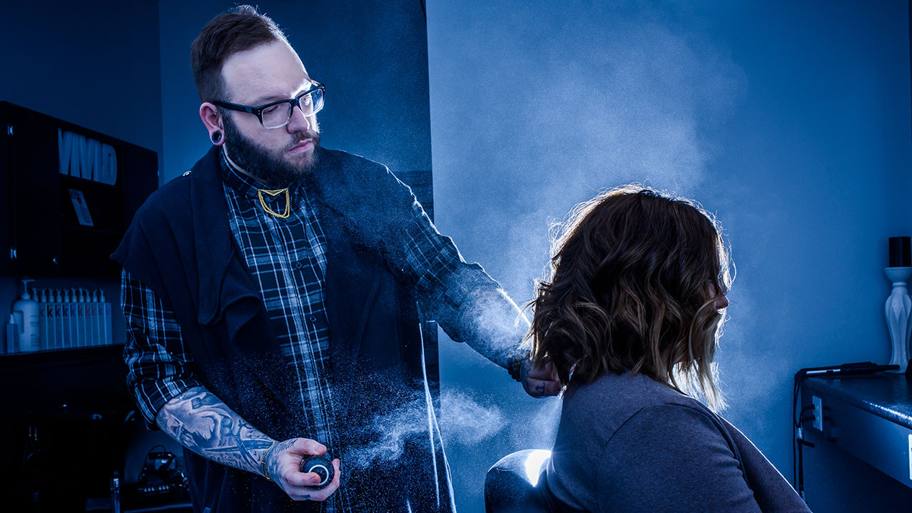 A stylist sprays hairspray on a client's freshly-cut hair in a salon setting.