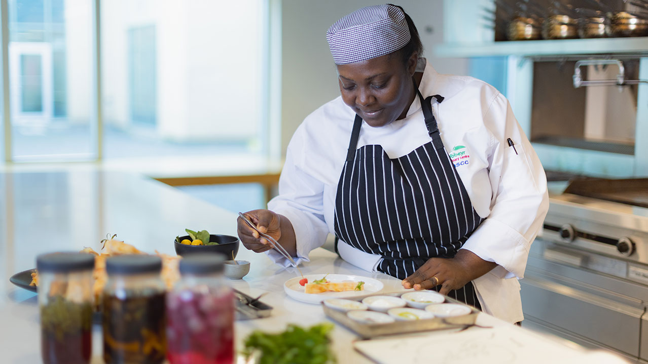 An NSCC student in a chef's hat and apron plates food.