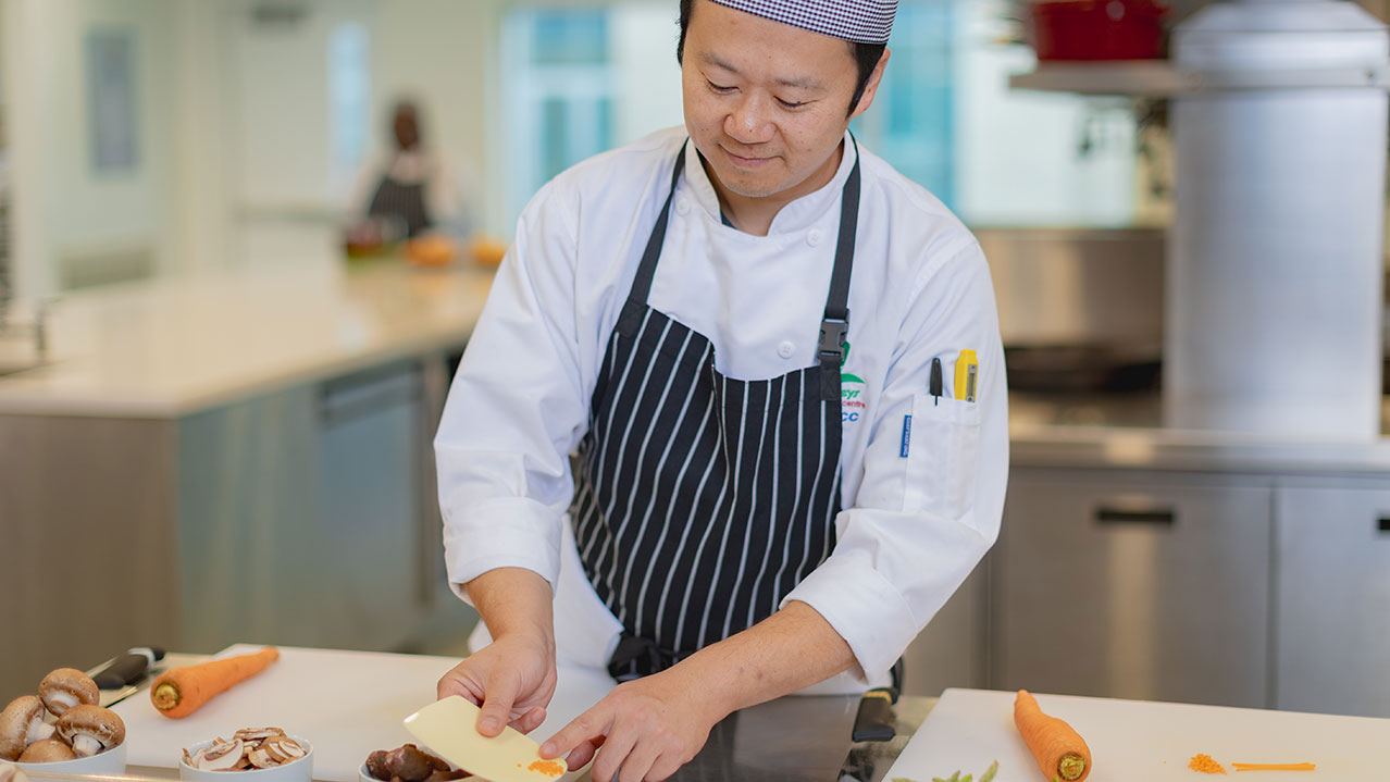 A Culinary Skills student wearing a chef's hat and uniform works in a kitchen preparing carrots and mushrooms.