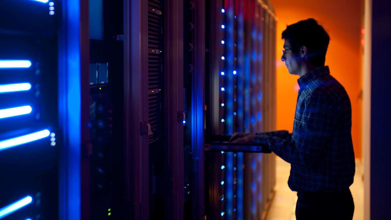 A man in a plaid shirt and glasses works in a computer server room.