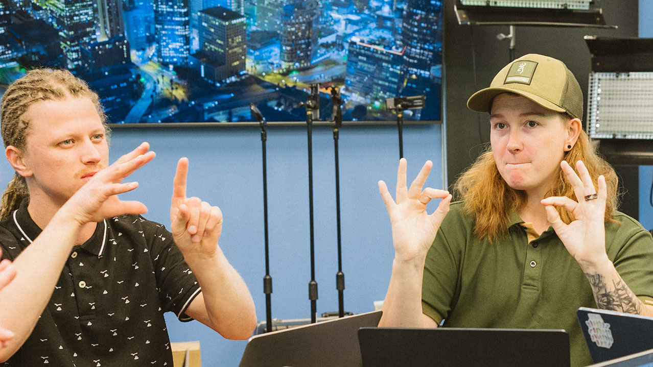 Two students are seated at a table signing to one another in American Sign Language (ASL).