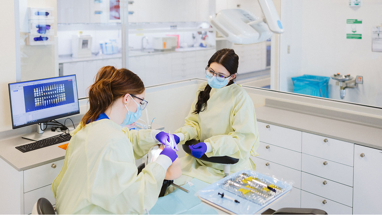 Two students wearing medical coats, masks and gloves work on a patient in a dental operatory for their Dental Assisting Level II program.