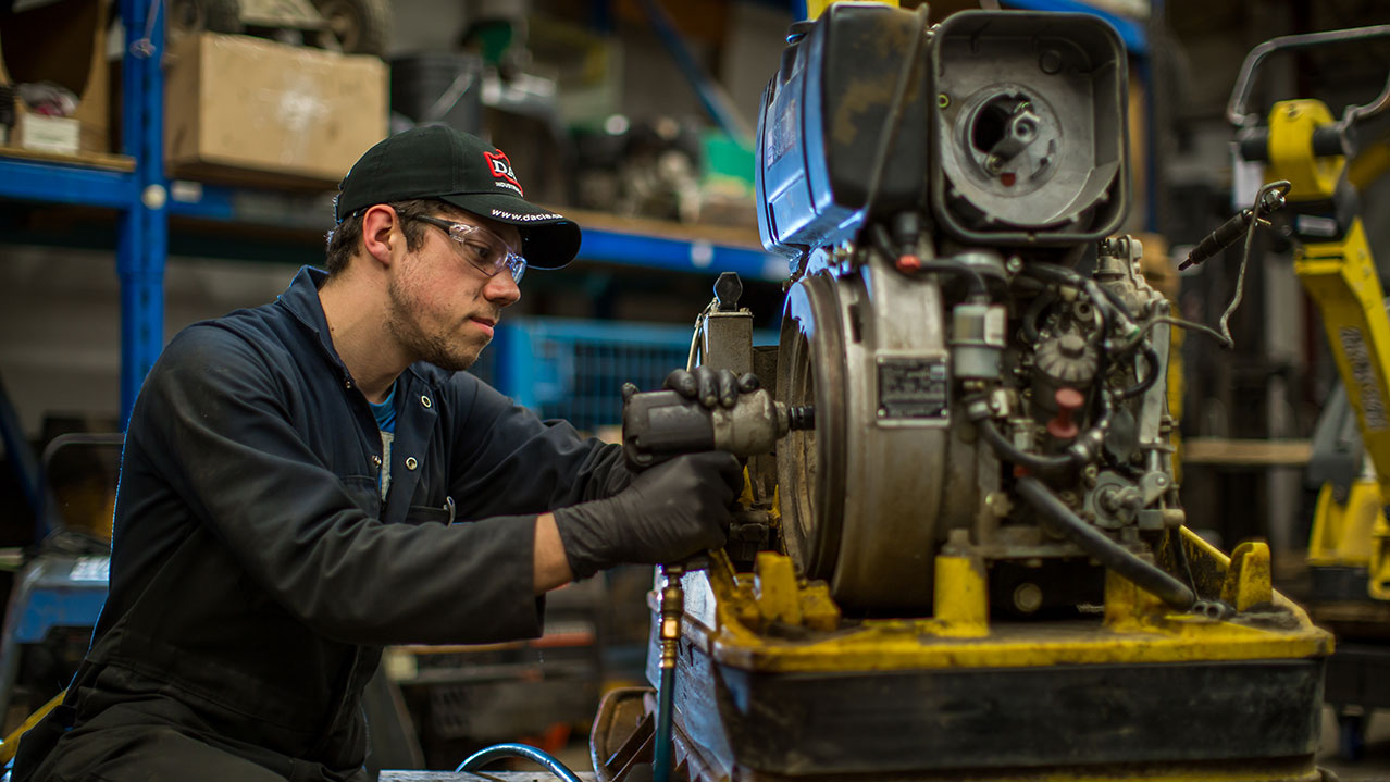 A man wearing a baseball hat, safety glasses and coveralls works on machinery in a workshop.