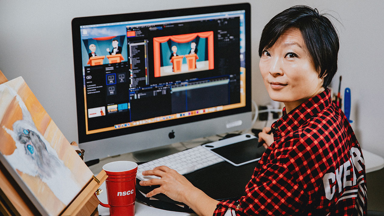 A student wearing a red and black checkered shirt sits at her computer and looks over her left shoulder while smiling at the camera.