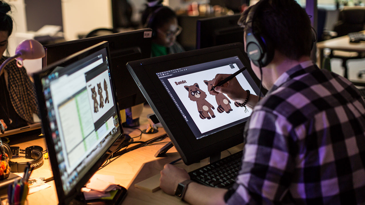 A person wearing a black and white plaid shirt and headphones leans over a tablet to create a digitally animated character.