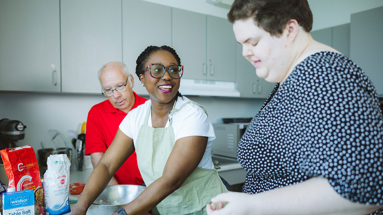 A disability support worker smiles while making cookies in a kitchen with two people she is working with.