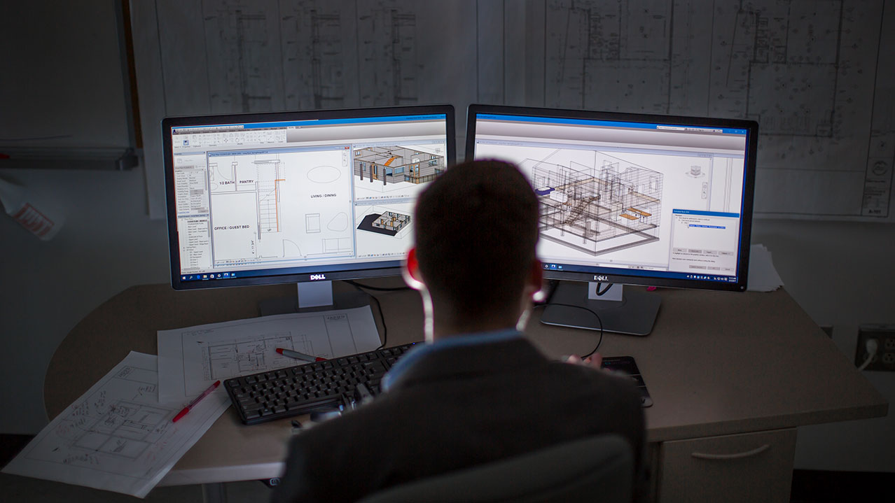 A man sits at a desk and looks at technical architecture drawings on two side-by-side computer monitors.