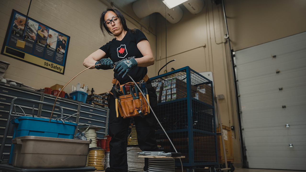 A woman standing in front of a work bench with many tools wearing safety glasses and gloves adjusts a thick, metal wire.