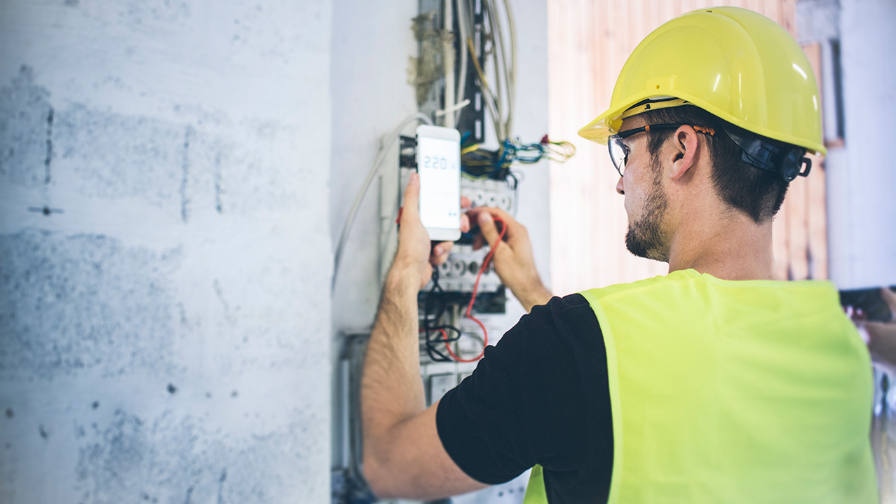 A man in a yellow hard hat, vest and safety googles uses equipment on the job for his industrial electrician course.
