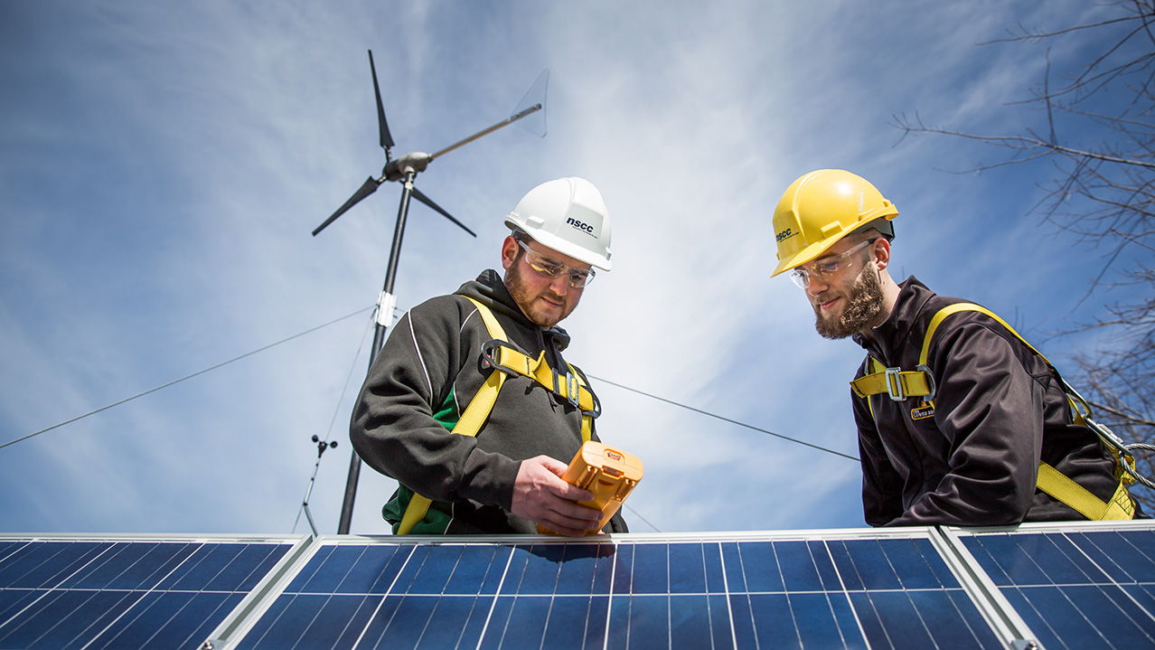 Two men in hard hats, safety goggles and safety harnesses hold a piece of equipment while standing near solar panels.