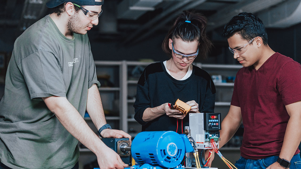 Three students in safety glasses learn about equipment in a campus workshop.