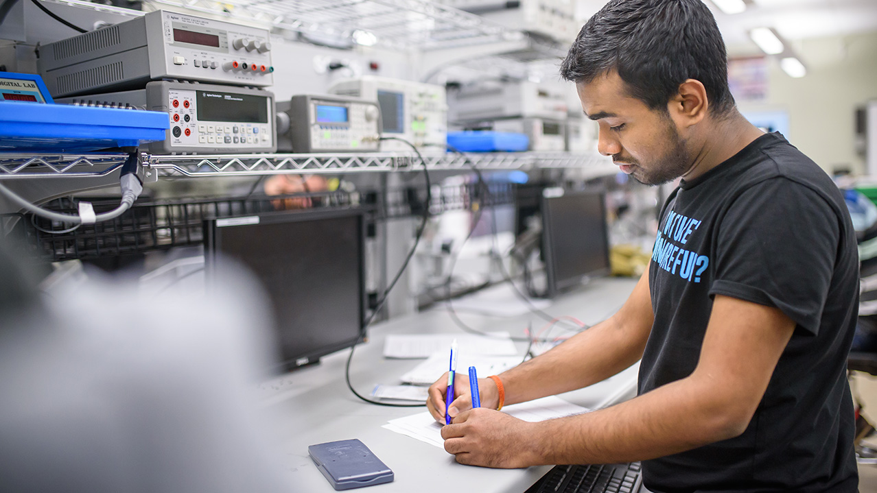 A student stands at a long desk covered in monitors and wires and other electronic equipment.