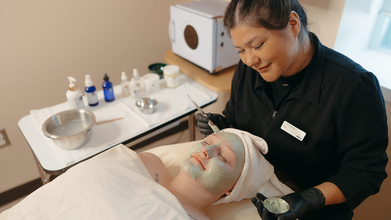 A smiling student practices giving a facial to someone in a spa setting.
