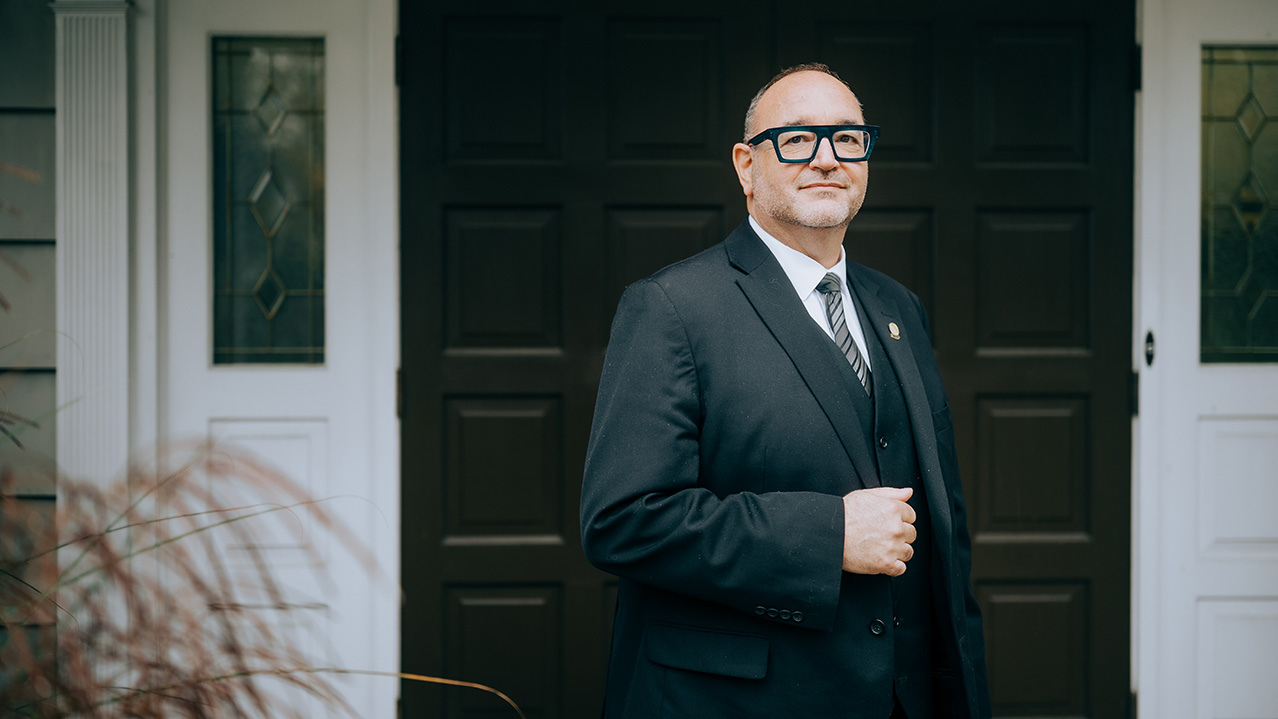A funeral director stands in a suit in front of a funeral home.