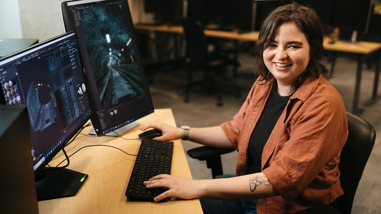 A smiling student in a computer lab sits at her computer with two large screens working on her interactive art.