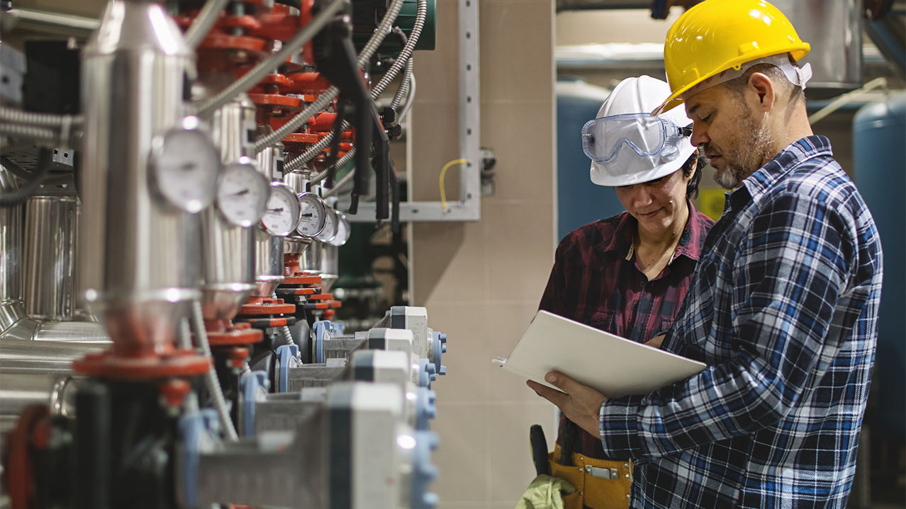 Two NSCC Gas Technician students wearing hard hats learn together in an industrial space.