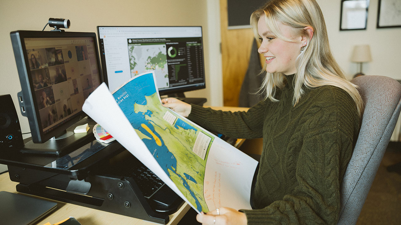 A student sits at a desk and smiles while looking at her computer screen and holding a paper map.