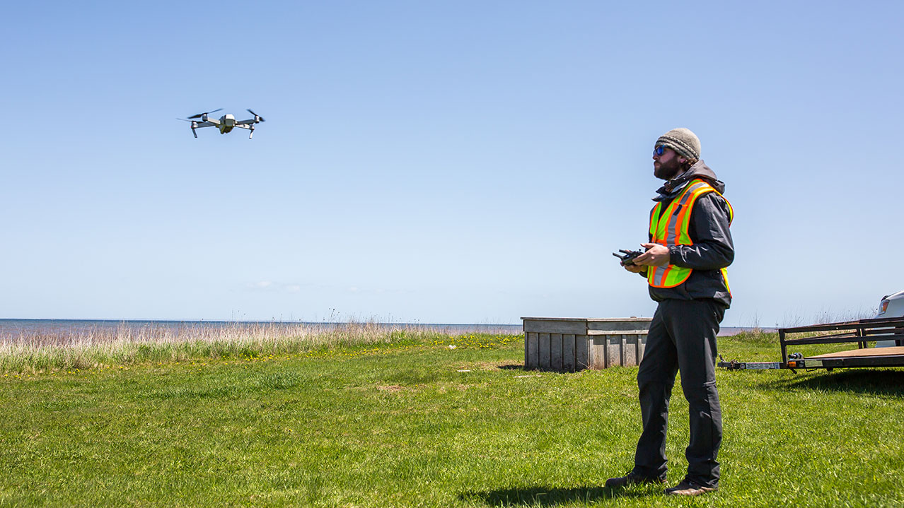A man in a safety vest stands in a field while operating a drone.