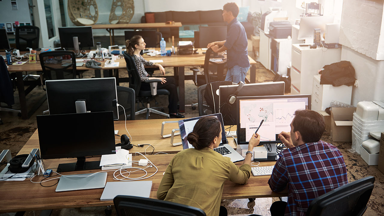 A man and woman in the geospatial analytics program look at a computer screen while two other people chat in the background.