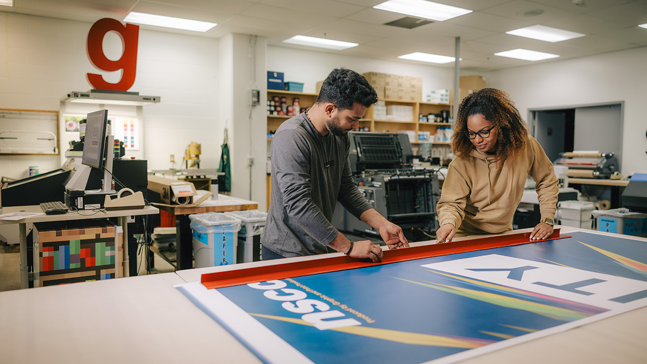 Two students work on a large printed poster in a print shop setting.
