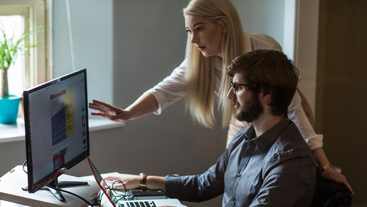 Two graphic design students look at a computer screen together with the male student with dark hair and glasses sits in front of the computer while the female student with long, blond hair stands beside him looking at the screen over his shoulder.