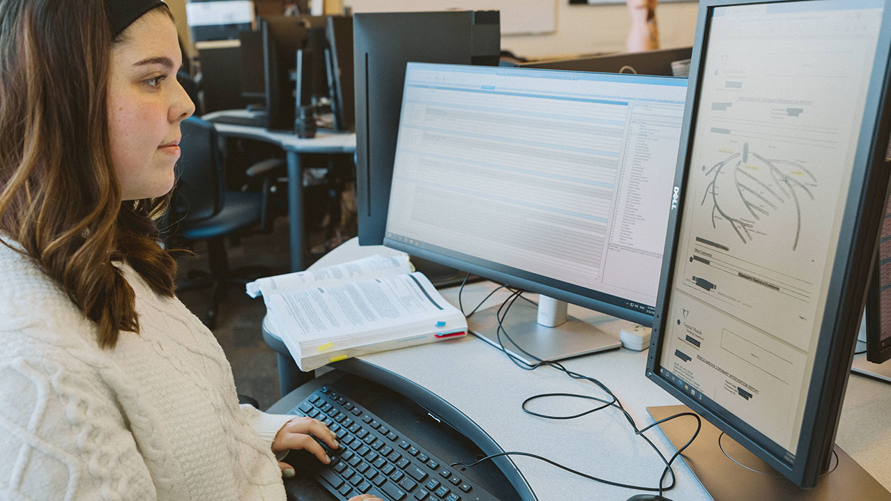 A student with long, brown hair and wearing a white sweater is seen sitting at a desk with her hands on the keyboard looking at 2 computer monitors for the health information management program. 