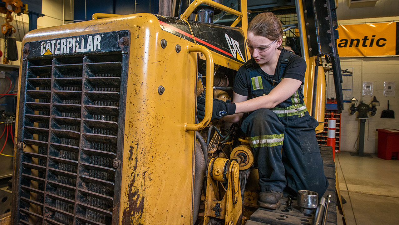 A woman in overalls works on a yellow tractor.