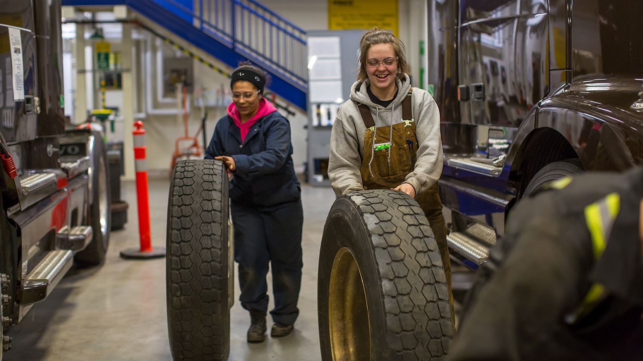 Two students in overalls roll large tires in NSCC's heavy duty equipment shop.