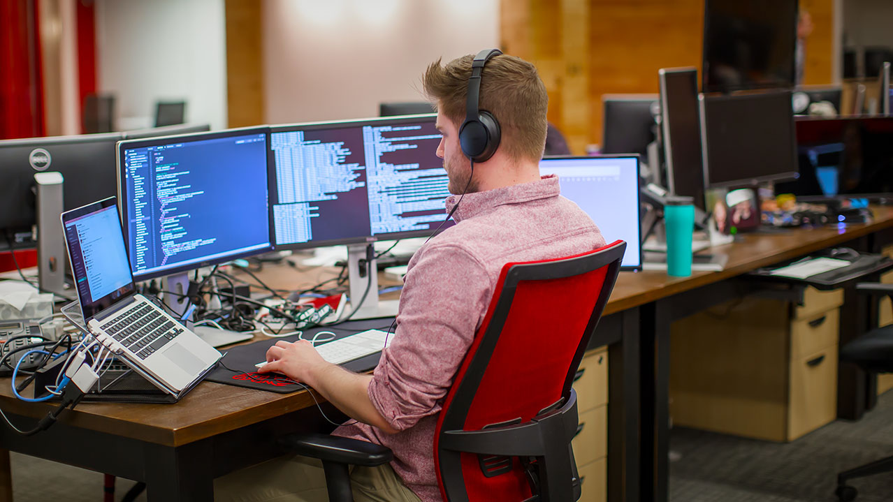 A man wearing headphones sits at a desk with five computer screens in front of him.