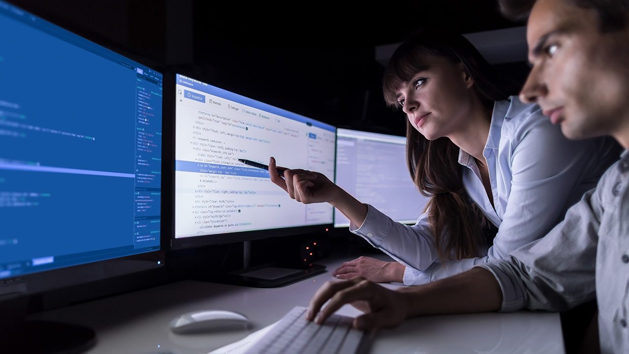 A man and woman gather around three computer screens; the woman points to one of the screens with her pen.
