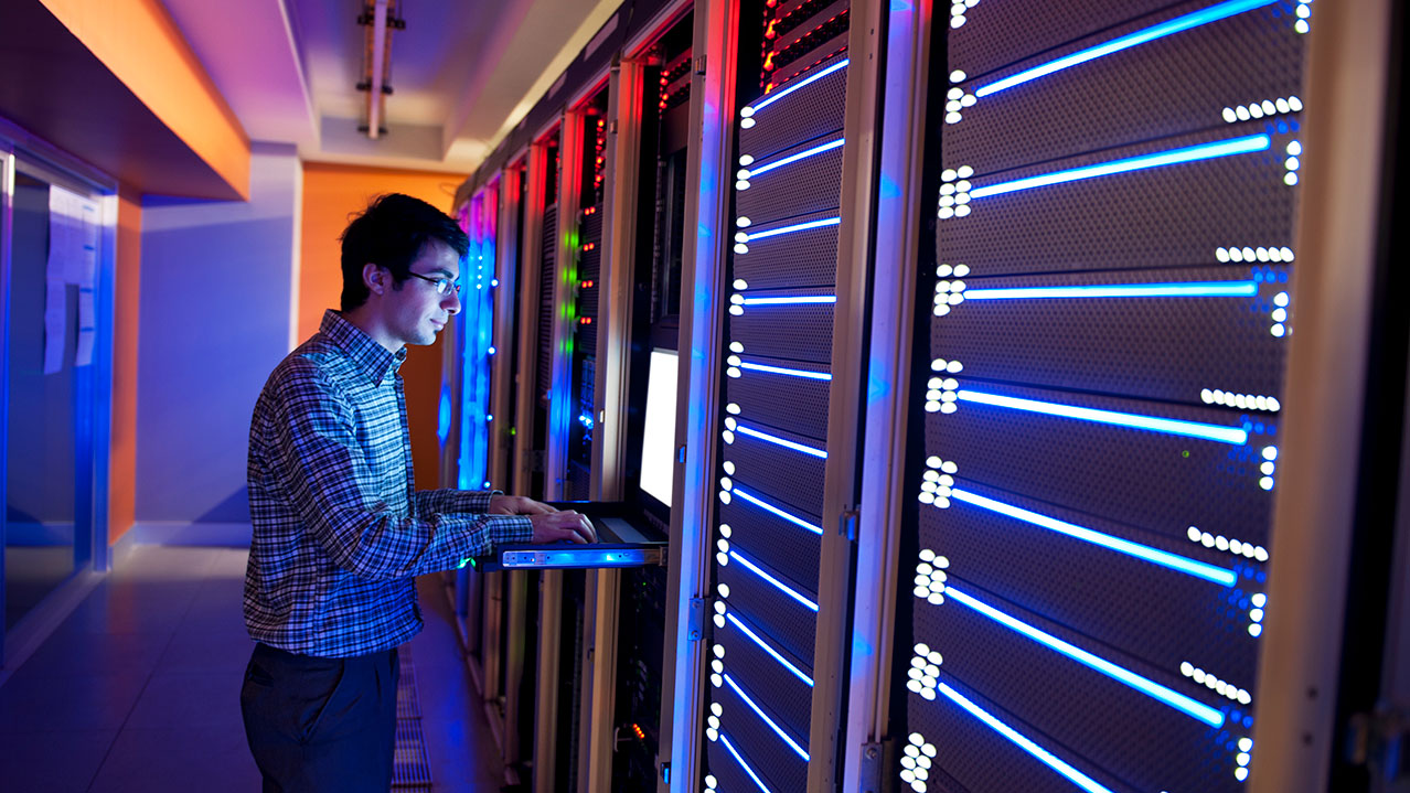 A man works on a computer while standing in a colourful computer server room.