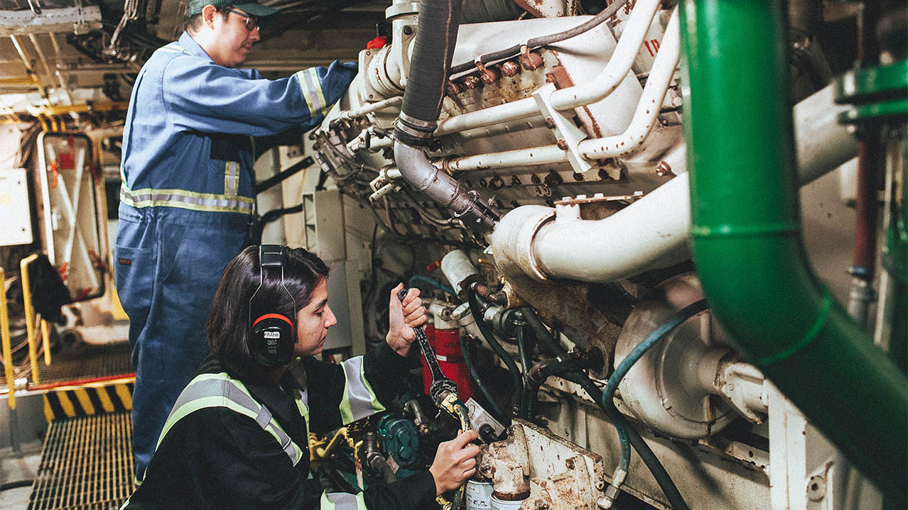 Two people, one wearing ear protection, work in a ship's engine room.