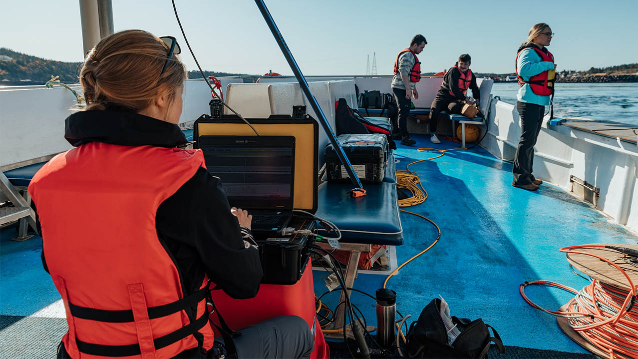 Four people, all wearing life jackets, work on the deck of a small boat; one woman has a laptop in front of her.