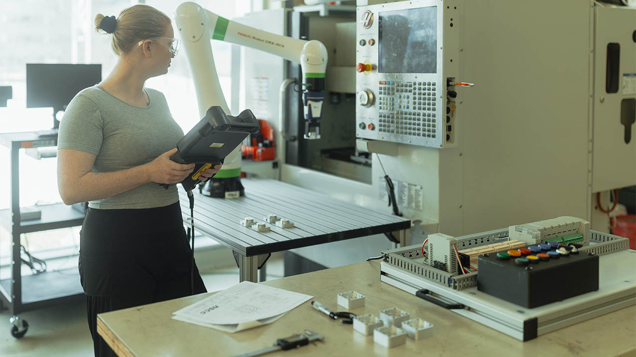 A woman in protective goggles using her tablet as reference, works on a robotic arm machine.