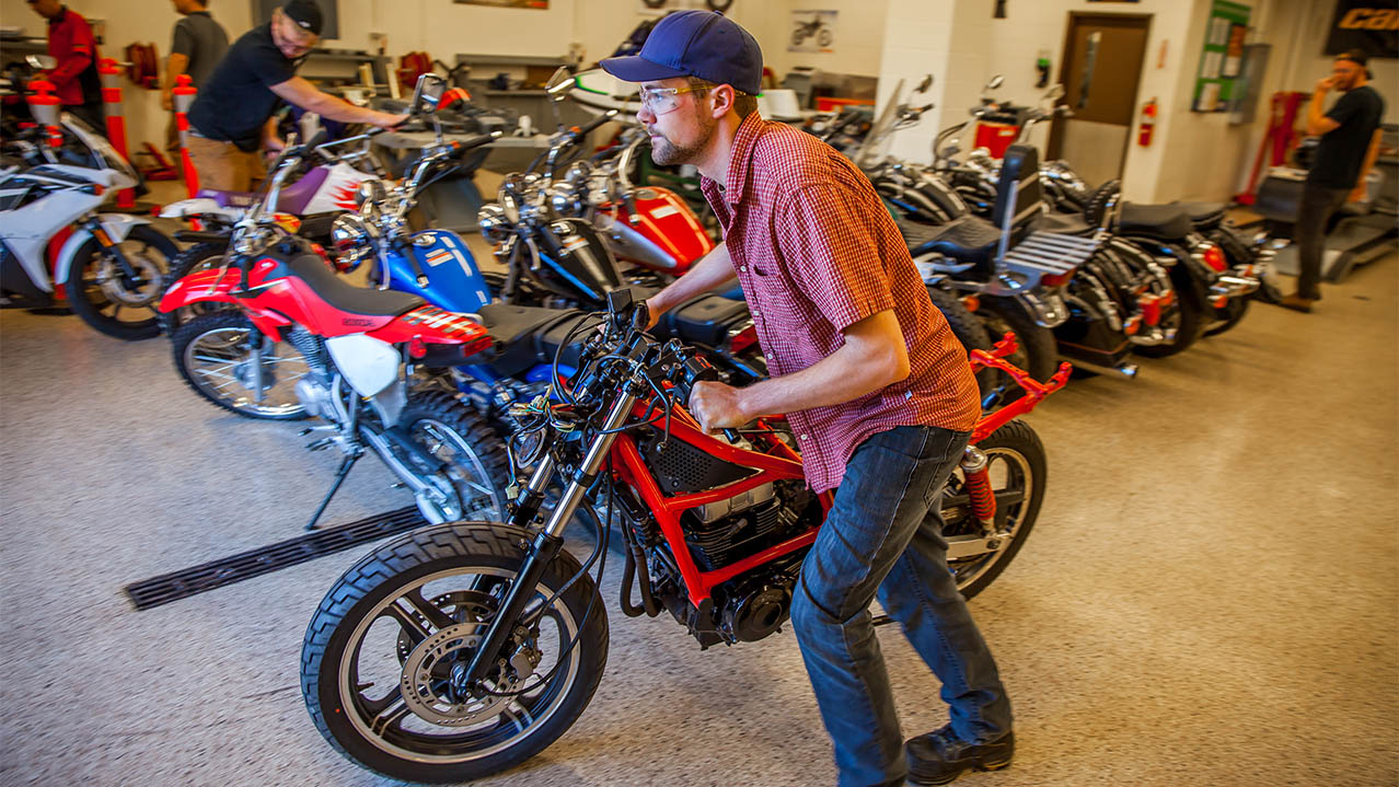 Students are seen in a garage filled with colourful motorcycles, moving them out of the garage to be worked on. 