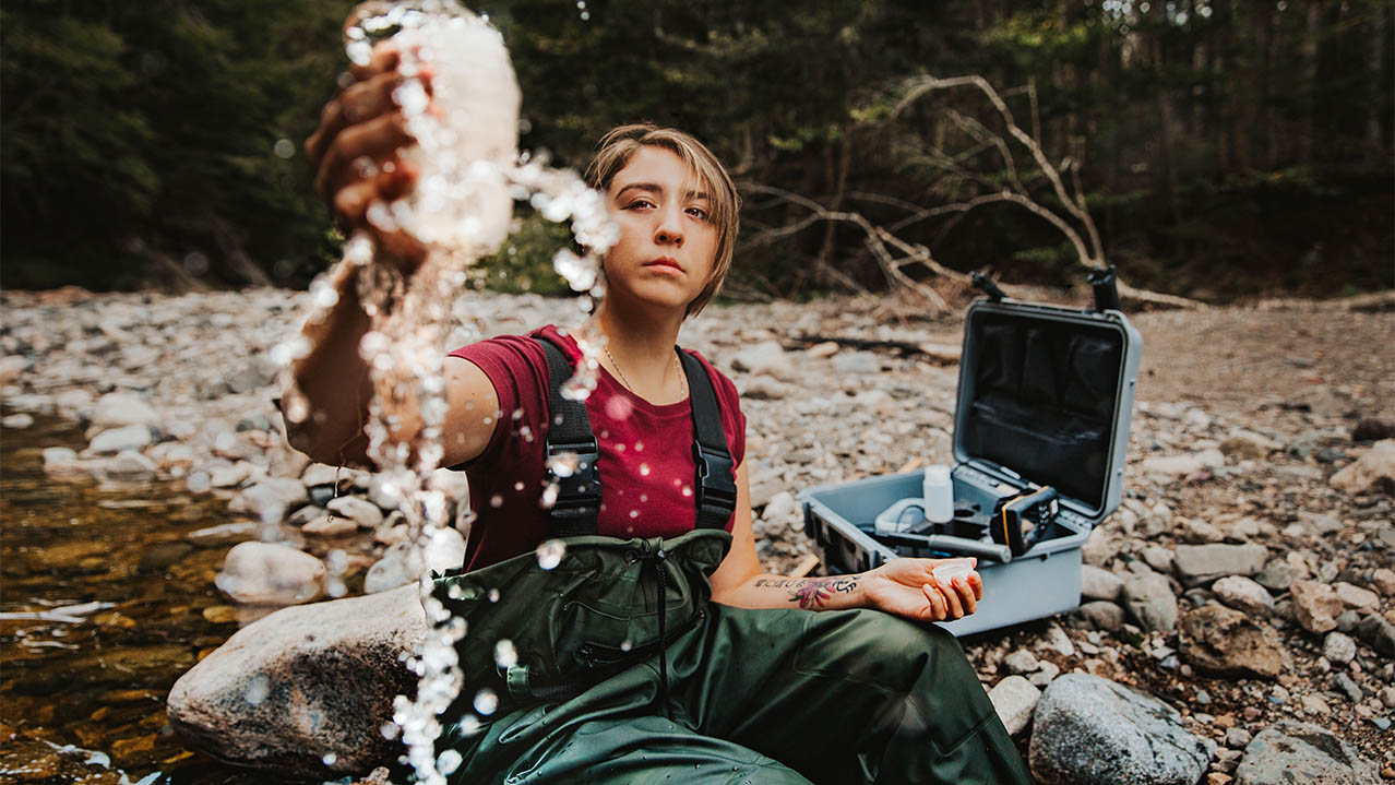 Kieran, a Natural Resources Environmental Technology program grad, sits on a rocky shoreline and fills a container with water