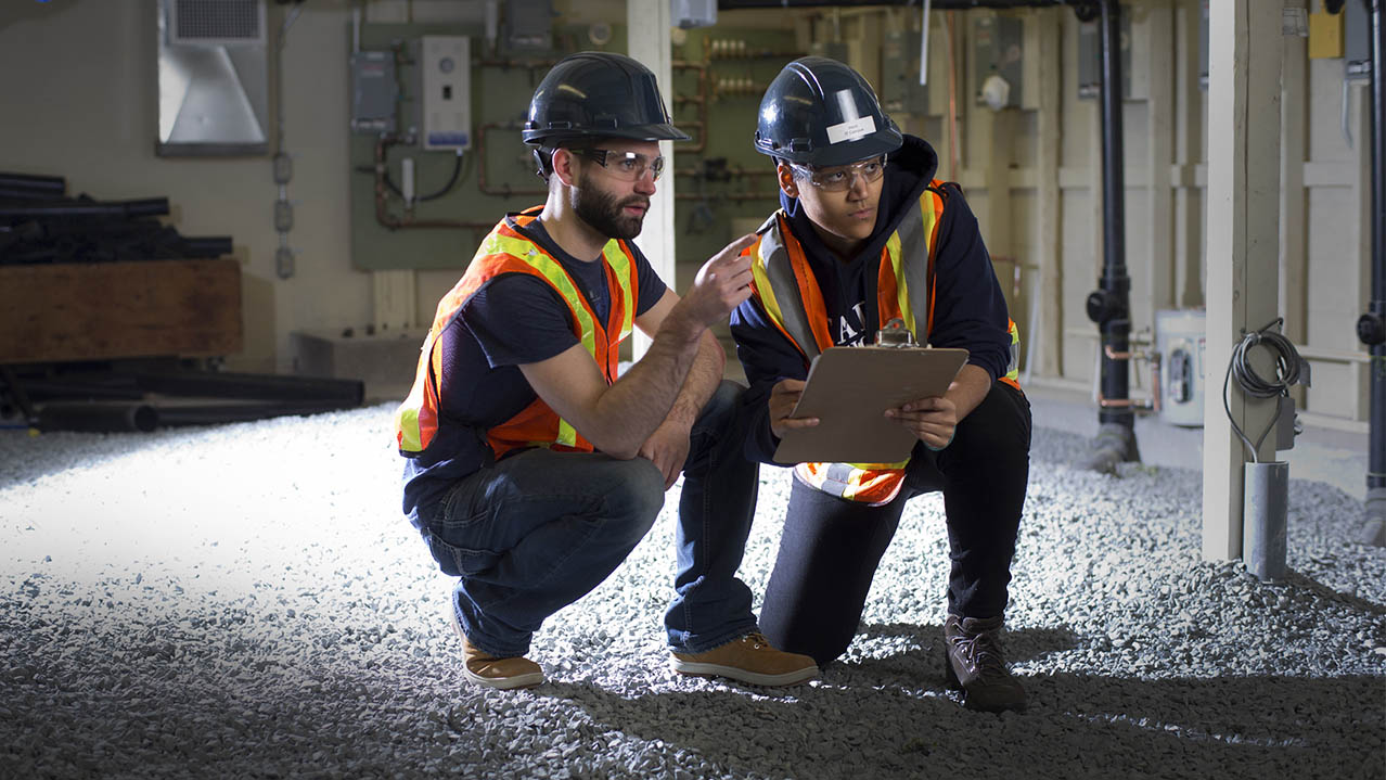 Two men in safety vests, goggles and hard hats consult a clipboard while kneeling in an unfinished basement or utility room.