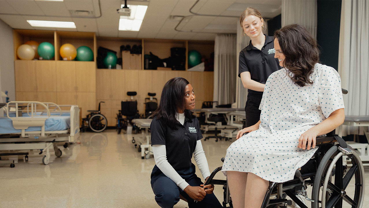 Two students wearing black shirt uniforms in a physiotherapy setting work with a patient sitting in a wheelchair.