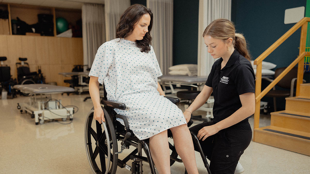 An Nova Scotia OTA-PTA program student kneels next to a patient in a wheelchair.