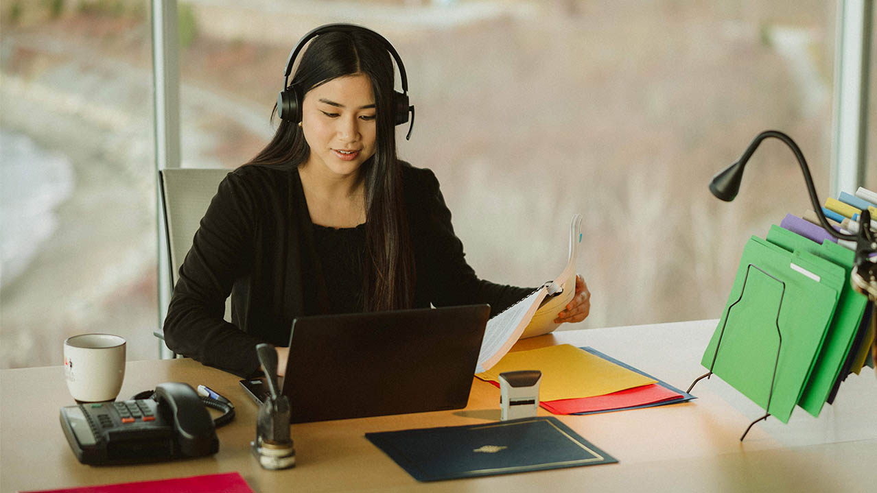 A woman with long, dark hair wearing headphones and sitting at a desk works on a laptop while holding a document.
