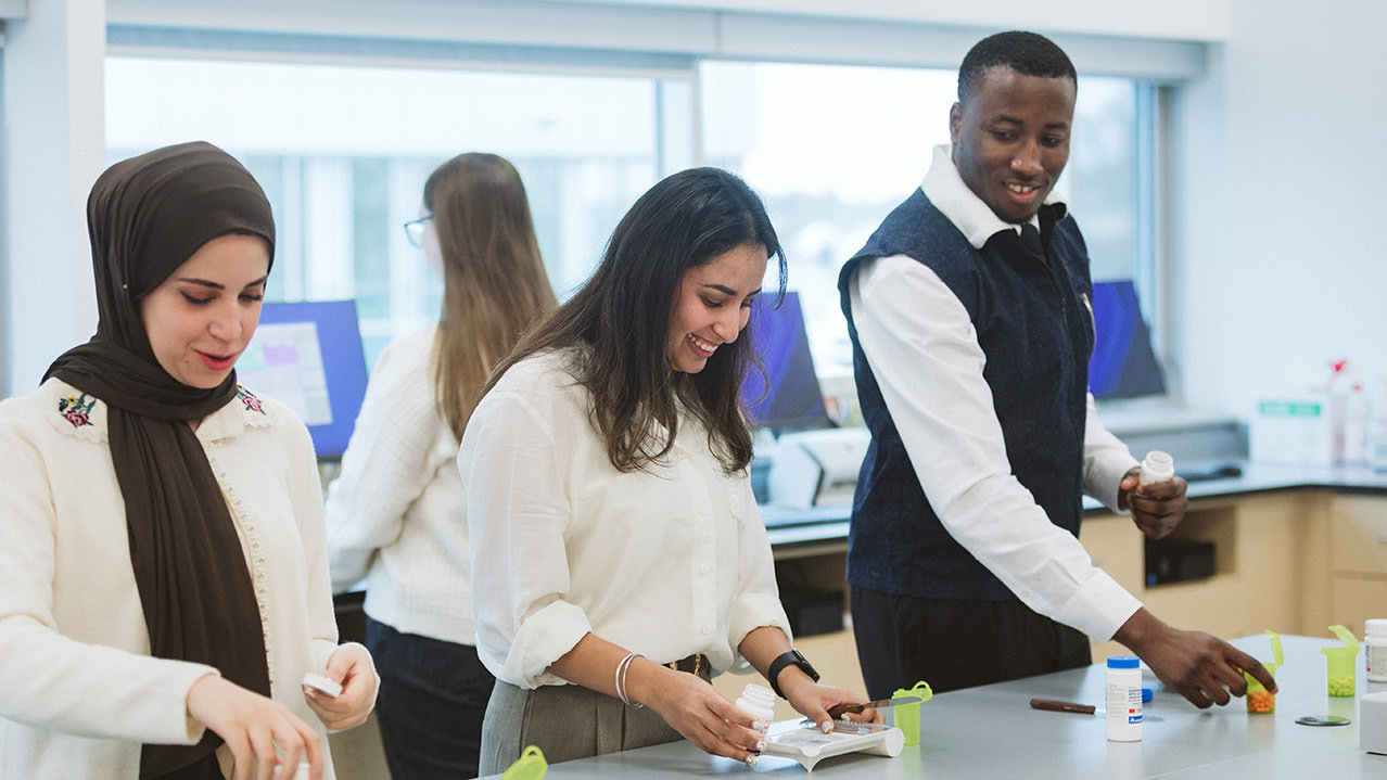 Three Pharmacy Technician students are seen sorting pills on a counter in a pharmacy classroom. 