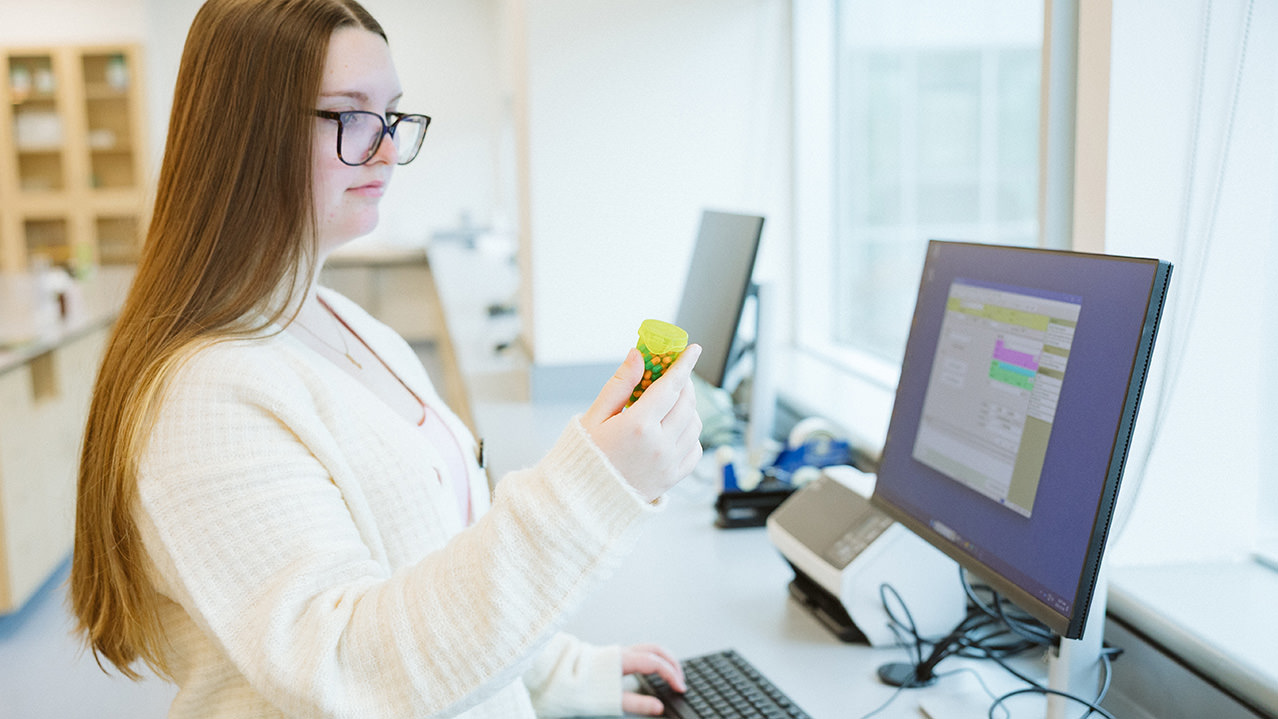 A female student wearing a white sweater is seen holding a bottle of pills and checking it matches what is on her computer.