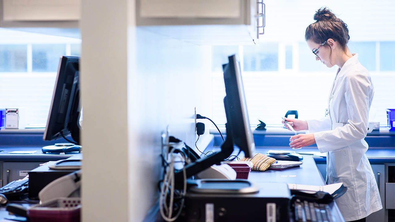 A woman in a lab coat works behind a prescription drop-off counter.