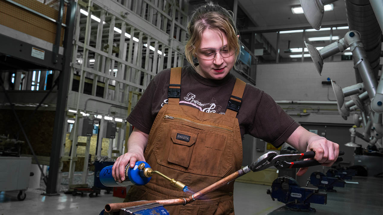A woman in brown overalls and safety glasses uses a blow torch on a piece of pipe.