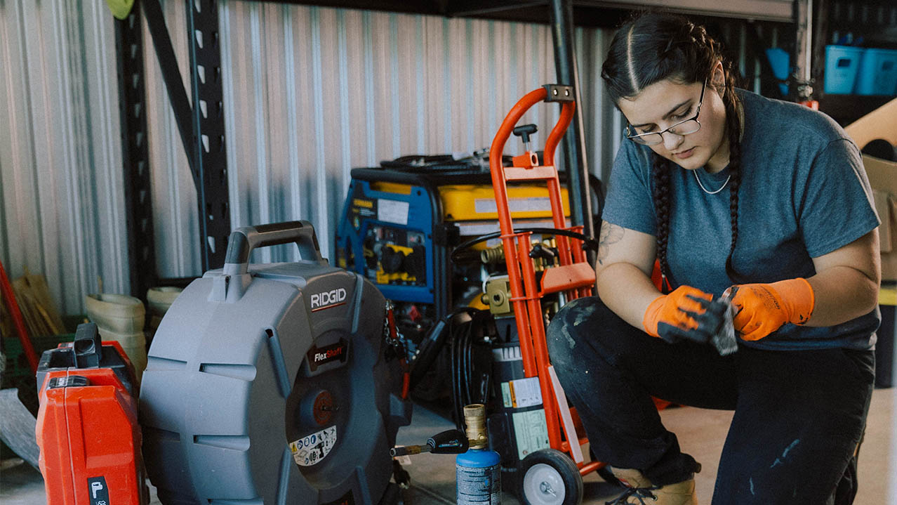 A woman wearing work boots, gloves and glasses, kneels on the floor next to equipment and works on some piping.